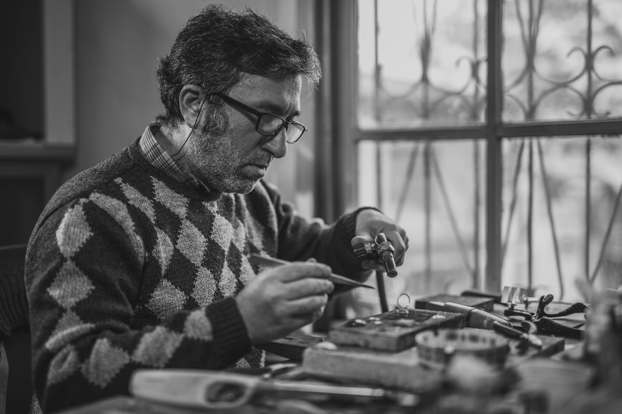 Black and white picture of a jeweler wearing glasses and a diamond-patterned sweater customising jewelry at a cluttered window-side workbench.