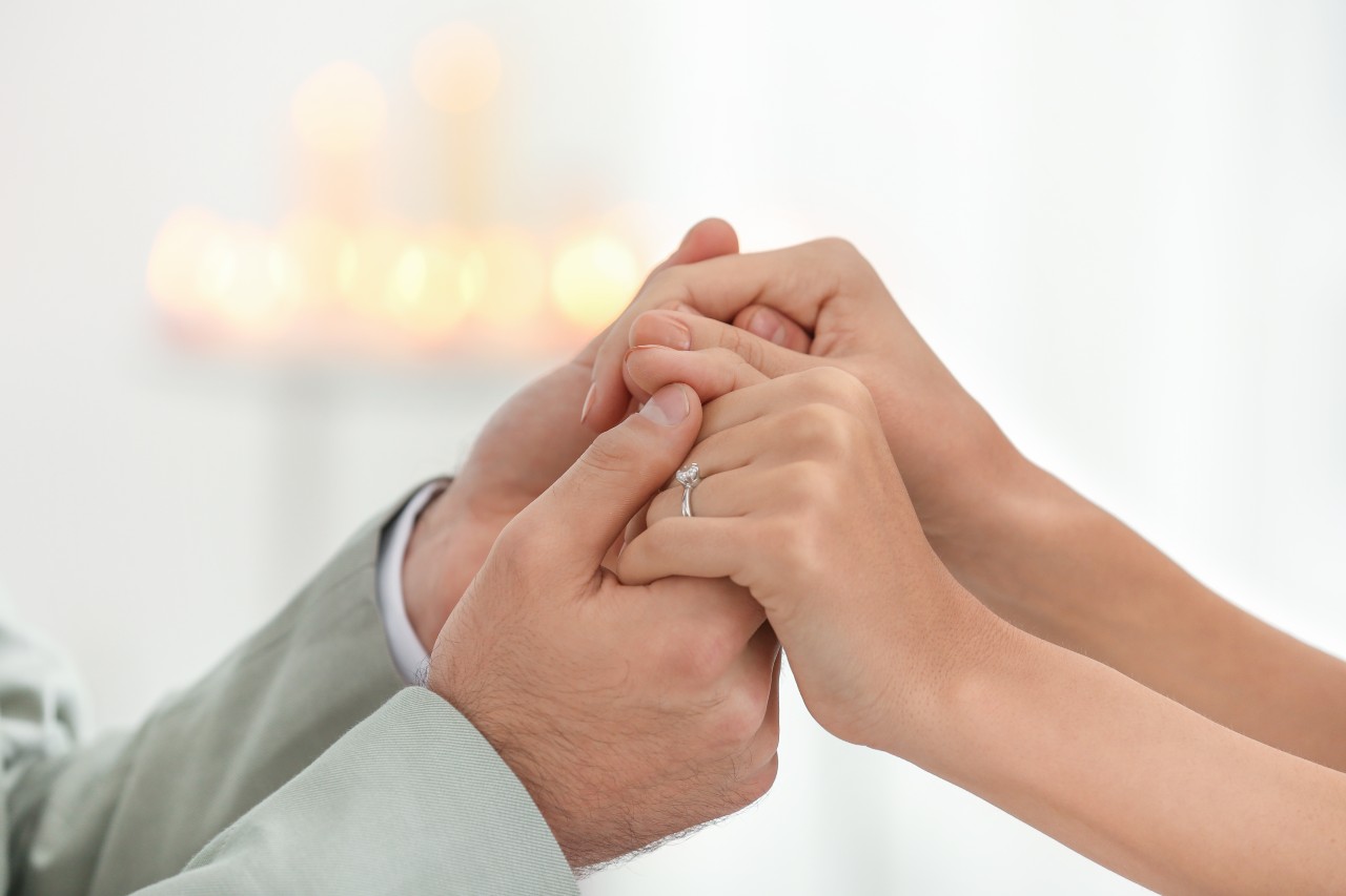Close-up of two people holding hands, one wearing a solitaire engagement ring, with a soft, blurred background of glowing candles.