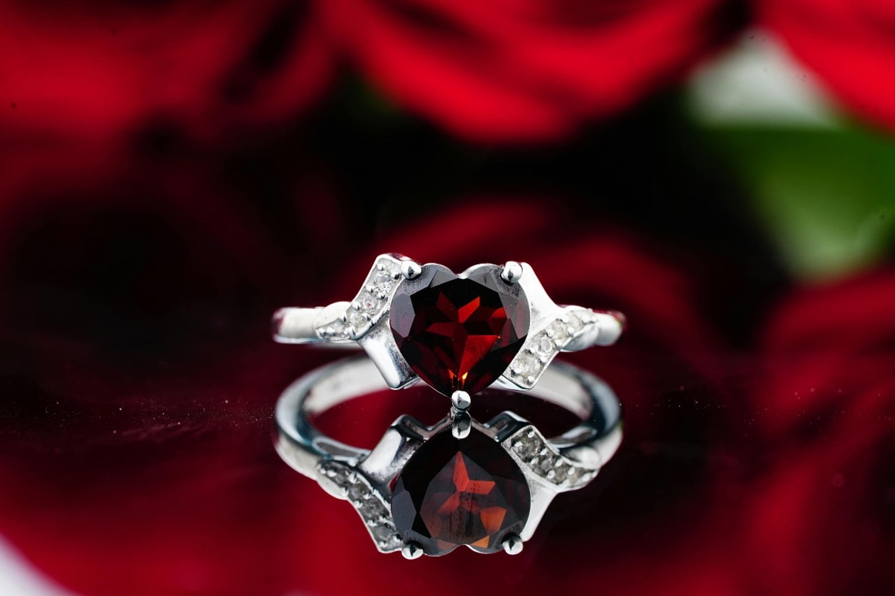 A silver ring with a heart-shaped deep red gemstone showcased against a blurry background of red roses